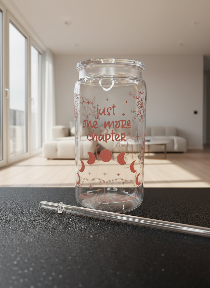 Clear glass tumbler with red text 'just one more chapter' and a metal straw on a black surface.
