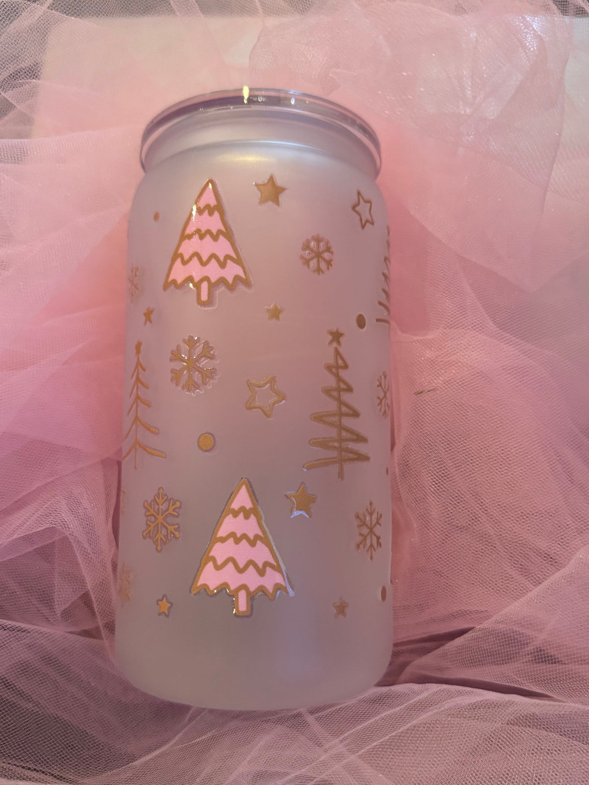 A frosted glass can cooler with a silver lid, decorated with pink and gold foil Christmas tree and snowflake designs, resting on pink tulle fabric.
