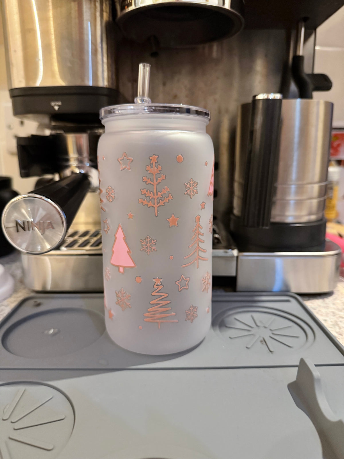 A frosted tumbler with a clear lid and straw, decorated with pink rose gold Christmas tree and snowflake patterns, sitting on a gray coffee maker drip tray.