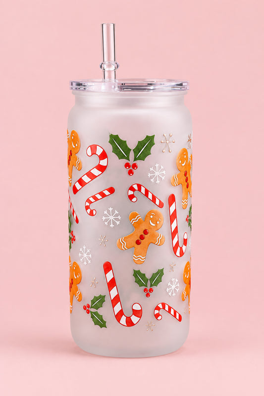 A frosted glass tumbler with a clear lid and straw, decorated with a festive gingerbread and candy cane pattern, sitting on a gray silicone mat on a kitchen countertop.