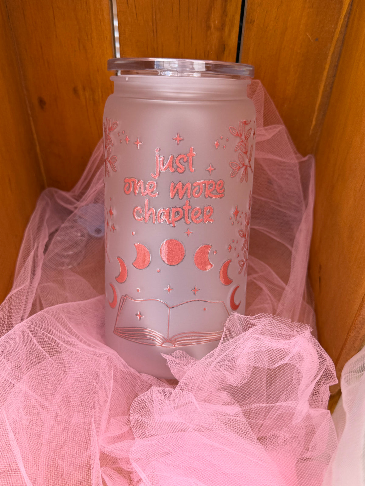 A frosted tumbler with a clear lid, decorated with pink celestial graphics and the text 'just one more chapter', resting on pink tulle fabric against a wooden background.