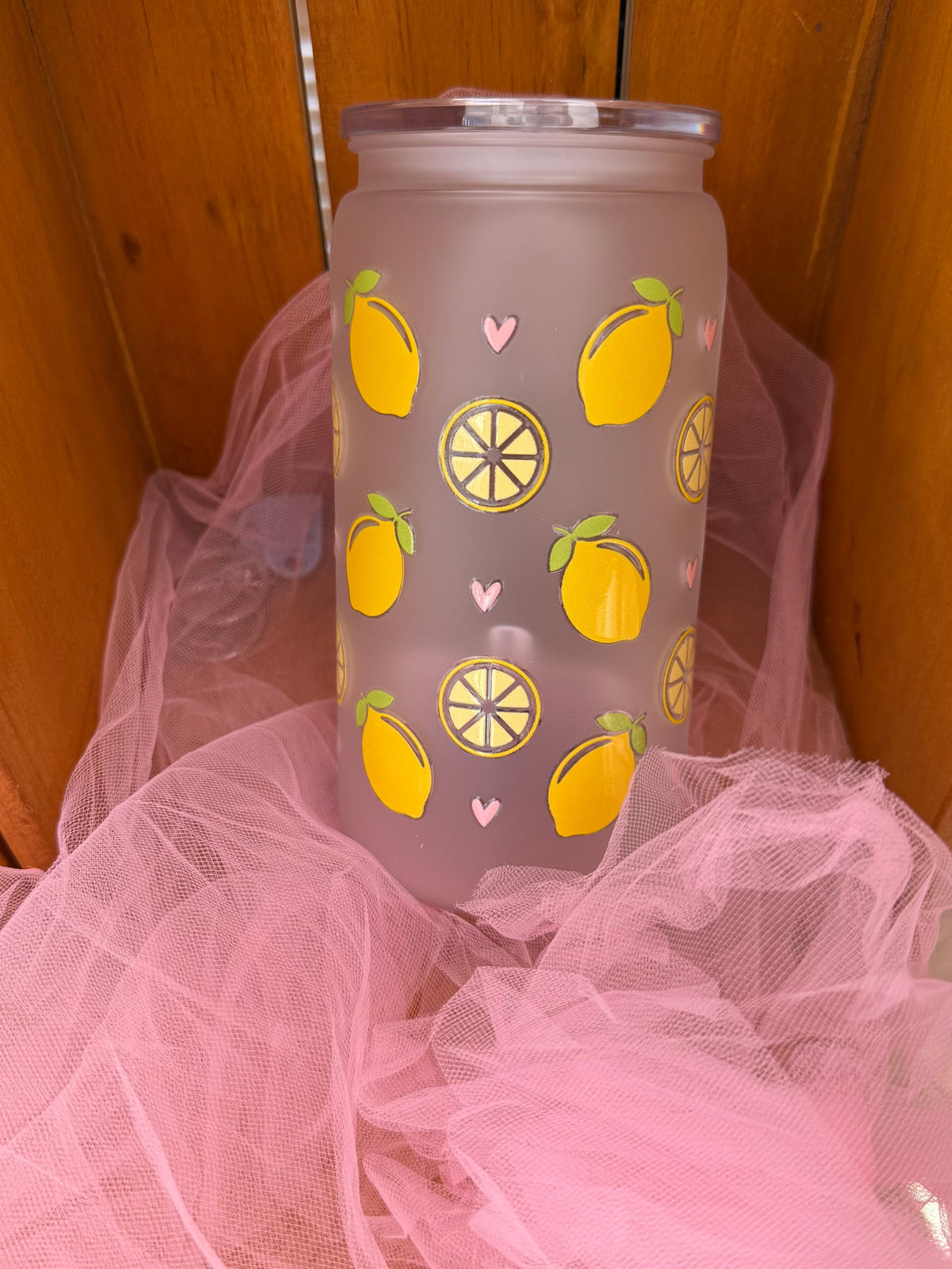 A frosted glass tumbler with a yellow lemon and lemon slice pattern, resting on pink tulle fabric inside a wooden box.