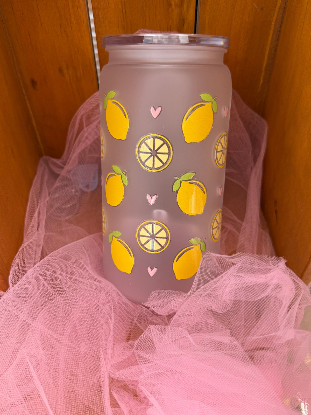 A frosted glass tumbler with a yellow lemon pattern, resting on pink tulle fabric inside a wooden box.